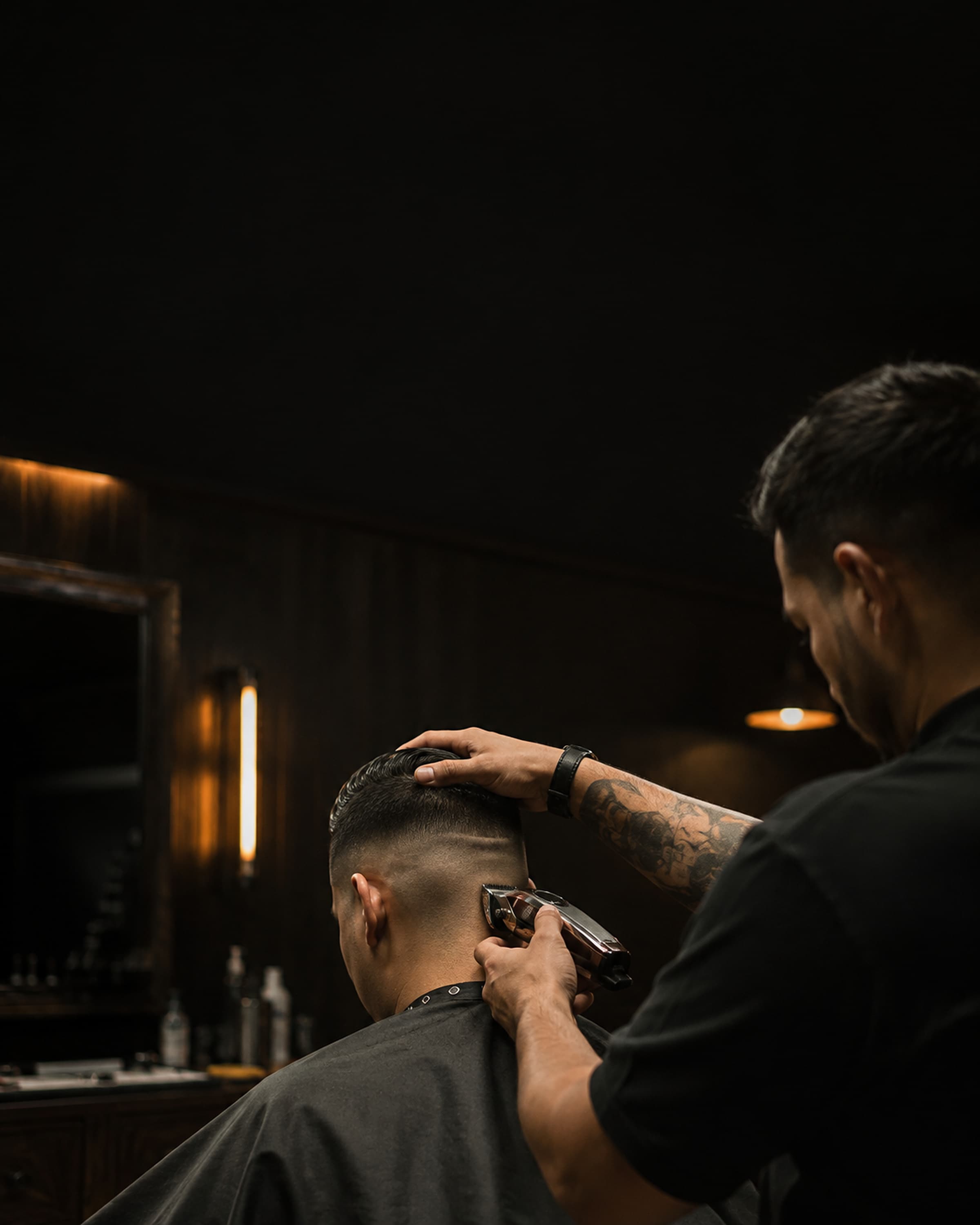 Barber mid-fade at the chair, clippers against the nape, shop lit low behind