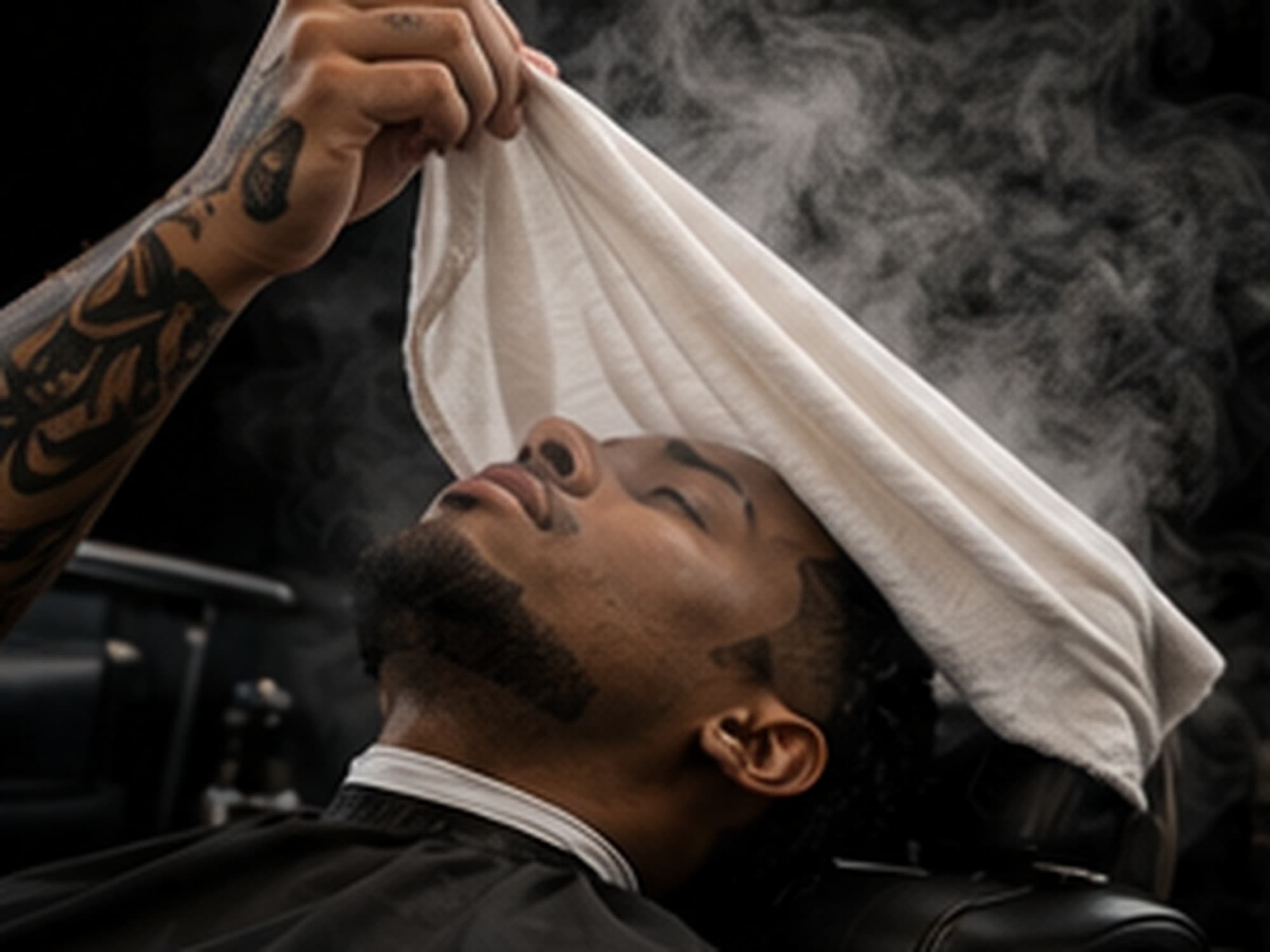 Hot towel lifted from a shaved jaw, steam visible against a dark background